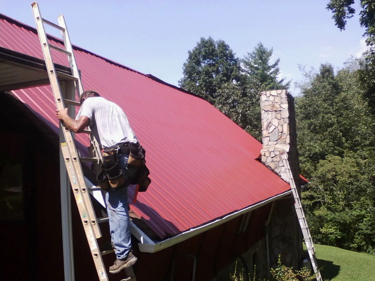 Roofer on a ladder beside a red metal roof and stone chimney — Soffit & Fascia Repair by Roofing Brothers in Rehoboth